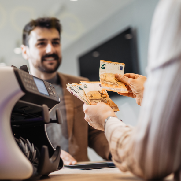 Mężczyzna uśmiechnięty z brodą i ciemnymi włosami stoi na wprost, a po drugiej stronie lady widać dłonie kobiety, która trzyma plik euro.